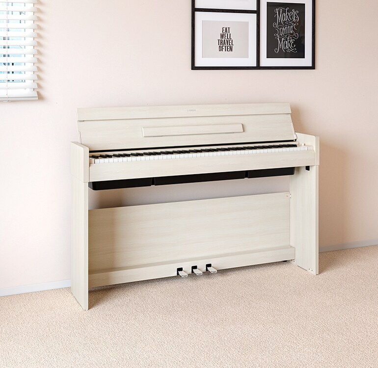 Ein Wohnzimmer mit einem ARIUS Piano in Weißer Birke, das neben Wandregalen mit Lampen und einem Diffusor steht und harmonisch in die Einrichtung integriert ist.
